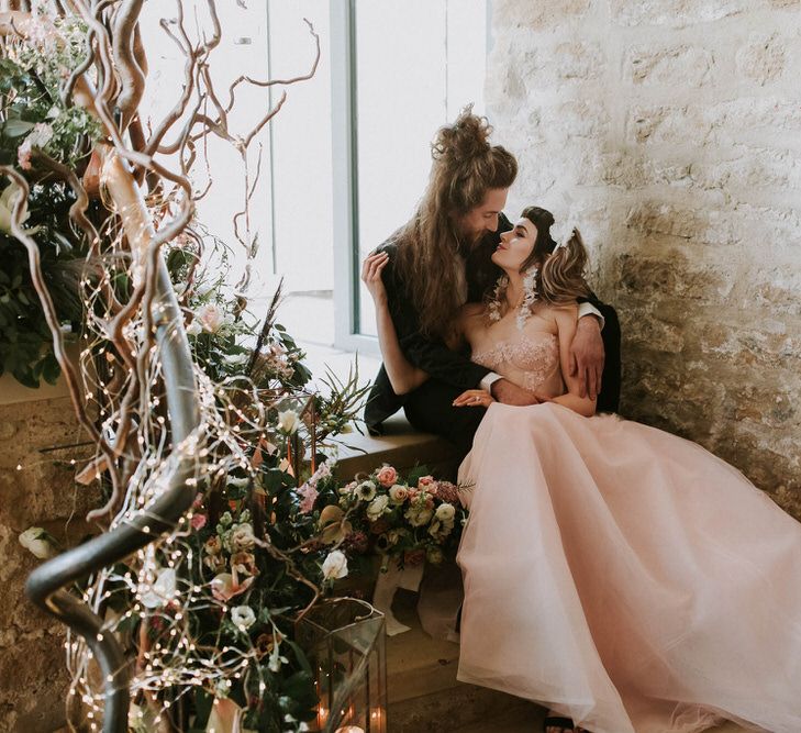 Bride in a pink strapless wedding dress sitting on the steps at Hooton Pagnell Tithe Barn with her groom in a black tuxedo 