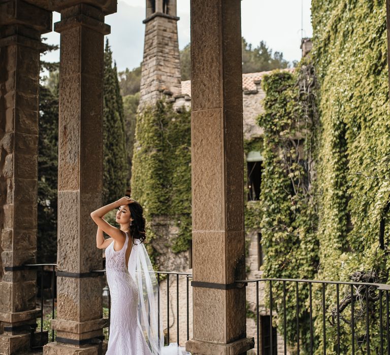 Bride stands beneath rustic pillars outdoors at La Baronia in front of lush greenery across castle 