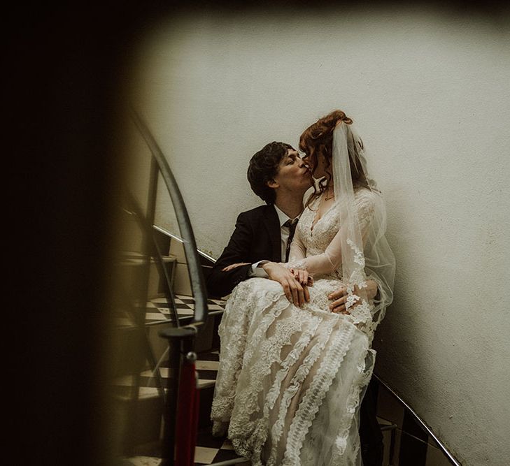 Bride wearing long sleeve lace wedding dress kisses her groom on staircase in moody styled picture at the Belair House