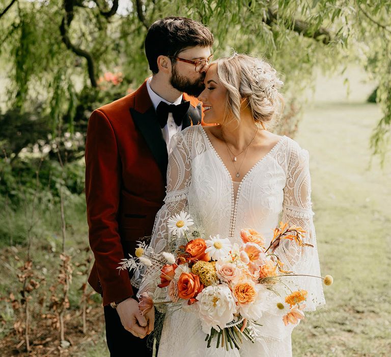 Groom in orange tux kisses the bride on the forehead in boho wedding dress with orange, pink and white bouquet 