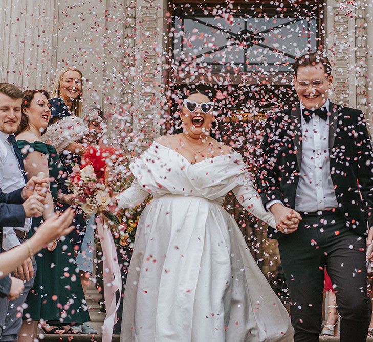 Bride wears off-the-shoulder ASOS curve wedding dress and carries bright floral bouquet as she walks through colourful confetti with her groom