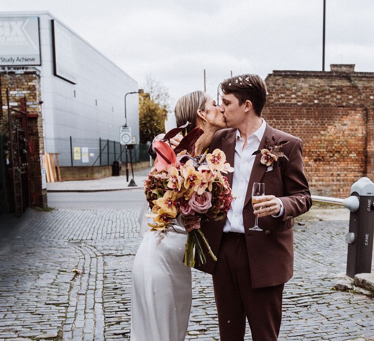 Groom in a brown suit with loose unbuttoned shirt kisses the bride in a satin wedding dress holding a pink and yellow bouquet 