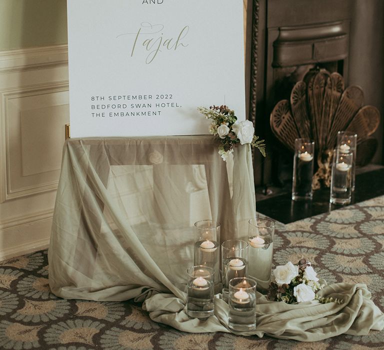 Minimal and simple wedding welcome sign with sheer green drapery, white flowers and floating tea lights in vases