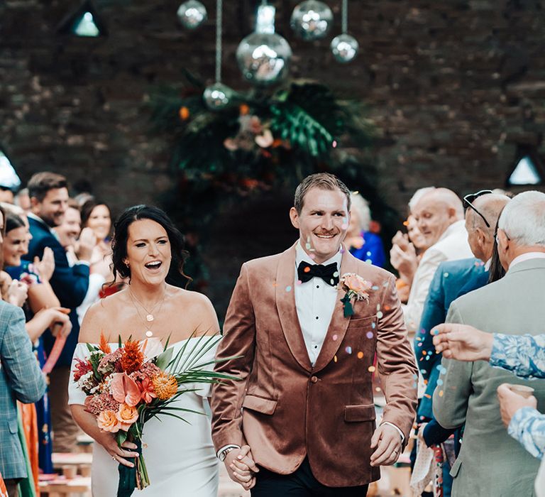 Bride in an off the shoulder wedding dress and the groom in a velvet pink jacket walk back down the aisle as a married couple