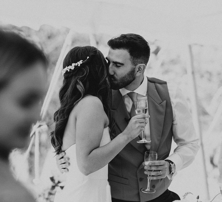 Groom in a light grey waistcoat and pale blue tie kisses the bride as they stand at their marquee wedding reception 