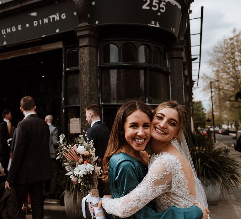 Bride in lace wedding dress hugs her friend outside pub reception 