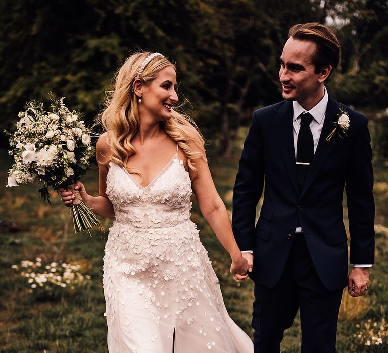 Bride wearing a pearl headband and pearl earrings in an applique wedding dress walks with the groom in a navy blue suit 