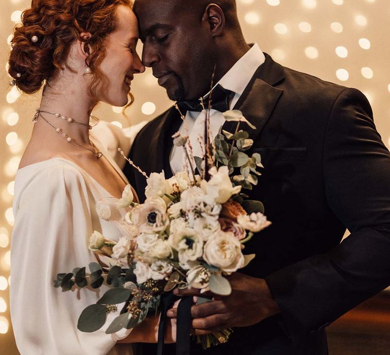 Bride in v neck satin wedding gown with double layered pearl necklace and pearl hair accessories holding minimalist floral bouquet embracing groom in black tux with fairy lights behind