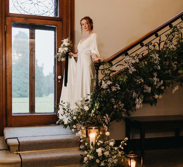 Bride in v neck satin wedding gown with double layered pearl necklace holding black and white neutral bouquet descending the stairs at Pepper Arden Hall