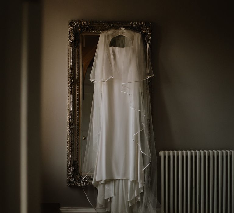 Square-neck satin wedding dress with full sleeves and large two tier veil hanging up on a mirror