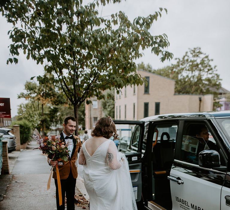 Groom holds Bride’s bouquet as she lifts her Dig For Victory Celestial wedding dress up to get into taxi