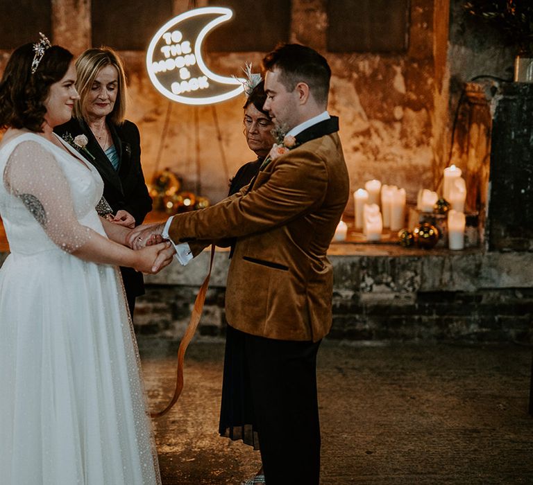 Bride and Groom take part in handfasting ceremony surrounded by candles and a moon neon sign for wedding lighting