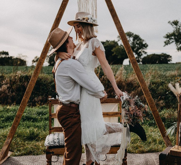 The groom in a boho wedding outfit and brown hat lifts up the bride who wears a boho wedding dress with Doc Martens 