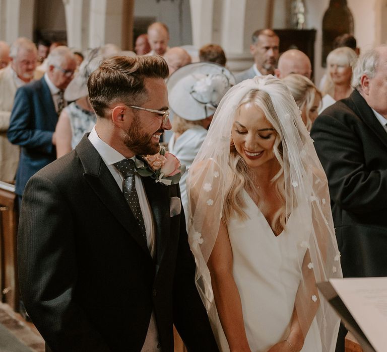 The bride wearing a flower applique veil and satin wedding dress smiling with the groom in three piece suit with large buttonhole for the church ceremony