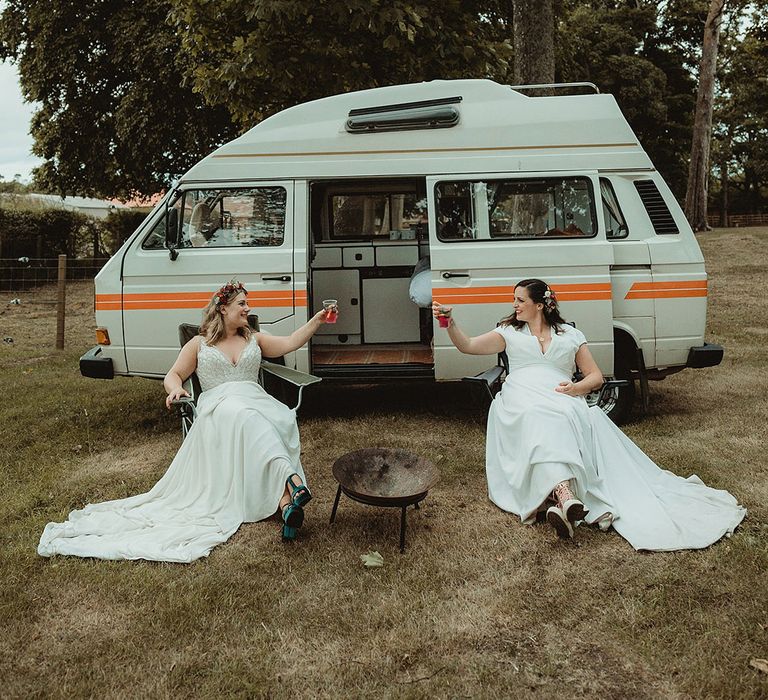 Brides sit outside their white and orange camper van on some folding chairs drinking 