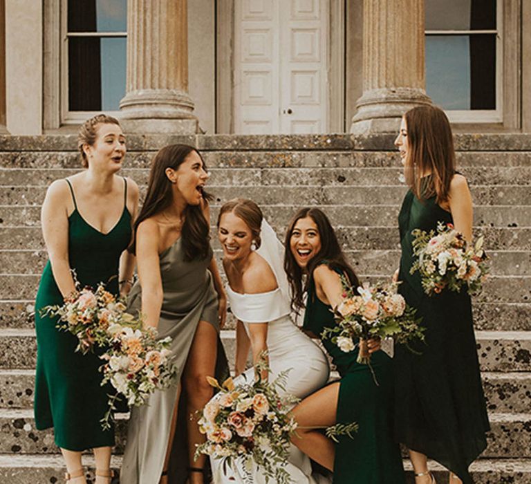 Bridesmaids all in satin dresses with mismatched colours laughing with the bride on the steps of the venue at The Grange