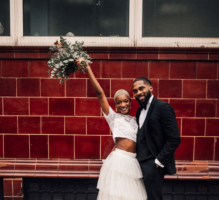 Black Bride and Groom pose with bridal bouquet in the air, featuring pink roses, eucalyptus and waxflower