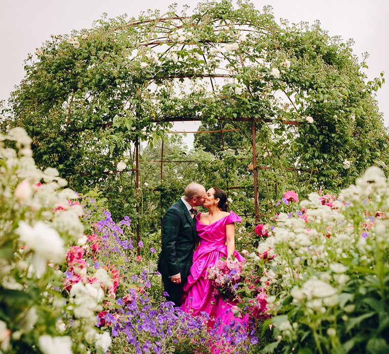 Bride in off the shoulder front ruching pink wedding dress kissing groom in dark forest green tartan suit with patterned tie, white pocket square and pink and green boutonniere surrounded by peonies, sweet peas, roses and baby's-breath florals at Middleton Lodge 