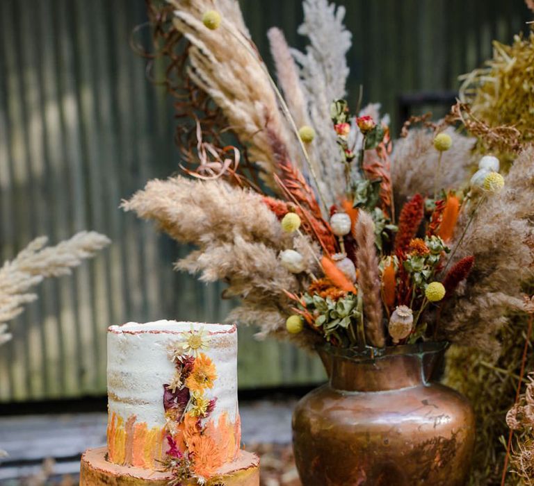 Three tiered vegan buttercream wedding cake in autumnal orange and cream ombre with dried floral decorations on the side next to large brass pot with autumnal dried flowers and pampas grass