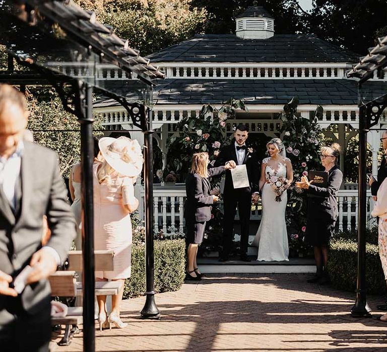 Bride in lace wedding dress with sheer, lace top and clusters of lace flowers on the shoulders and plunge neckline standing with groom in classic black tux with black bowtie and blush rose boutonniere at the bandstand at Old Kent Barn 