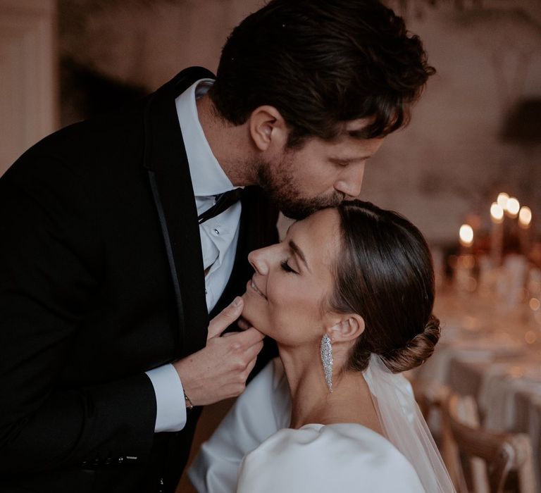 Groom in classic black tuxedo kissing bride on the head wearing puff sleeve wedding dress with church length veil and diamond drop earrings 