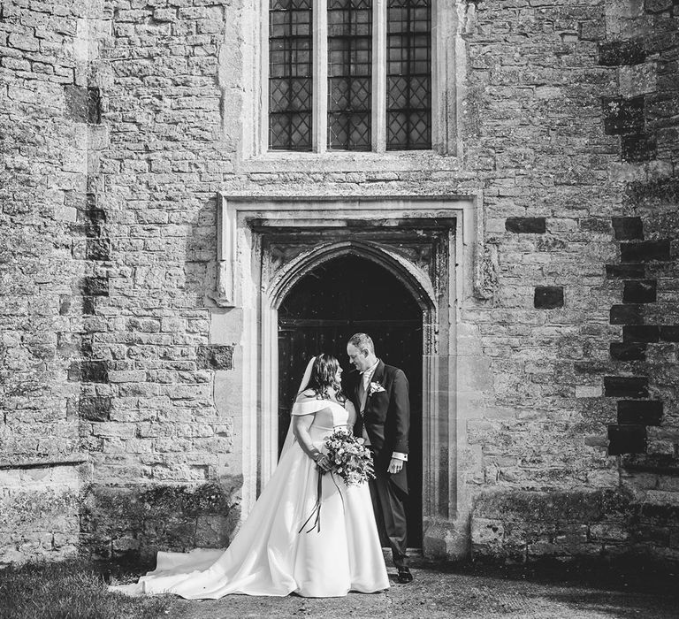 Groom in morning suit looks lovingly into the bride's eyes as she stands in a off the shoulder wedding dress outside of the church wedding 