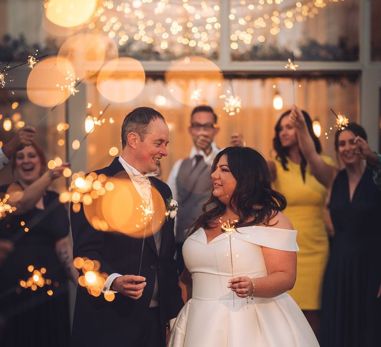 Groom in navy morning suit with white rose buttonhole and white tie with the bride in an off the shoulder wedding dress having sparklers for their sparkler send off 