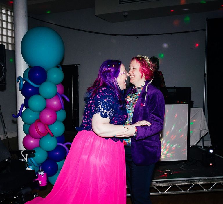 Bride in long pink wedding dress with purple sparkly bridal cover up embracing bride in purple velvet blazer and black suit trousers with purple bowtie, pink patterned pocket square and colourful patterned shirt