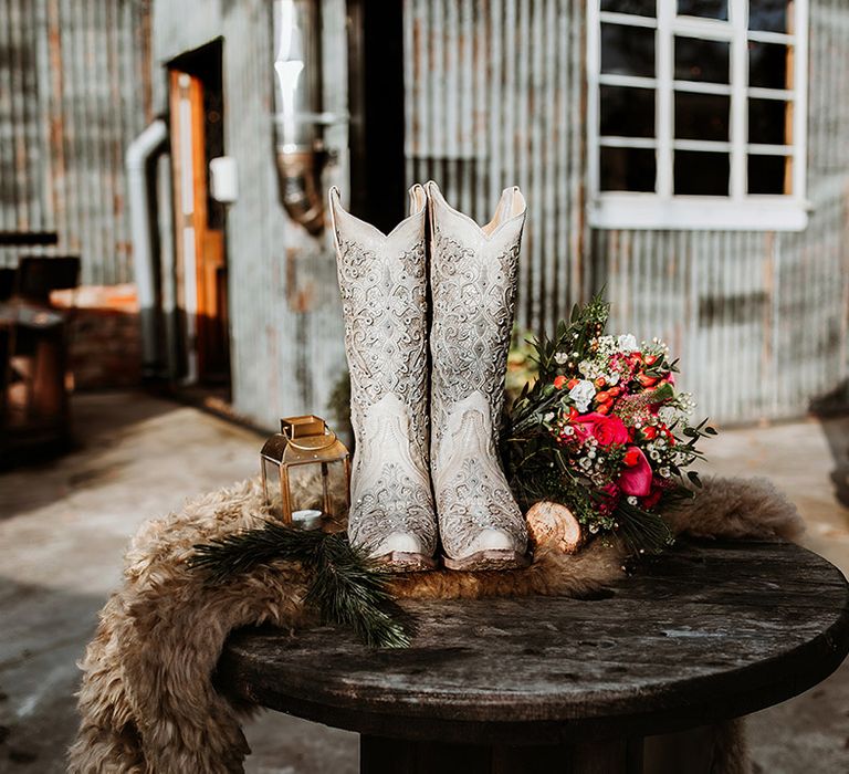 White bridal cowboy boots with silver glittery detailing on table with faux fur throw, tea light candle in brass lantern and colourful floral bouquet 