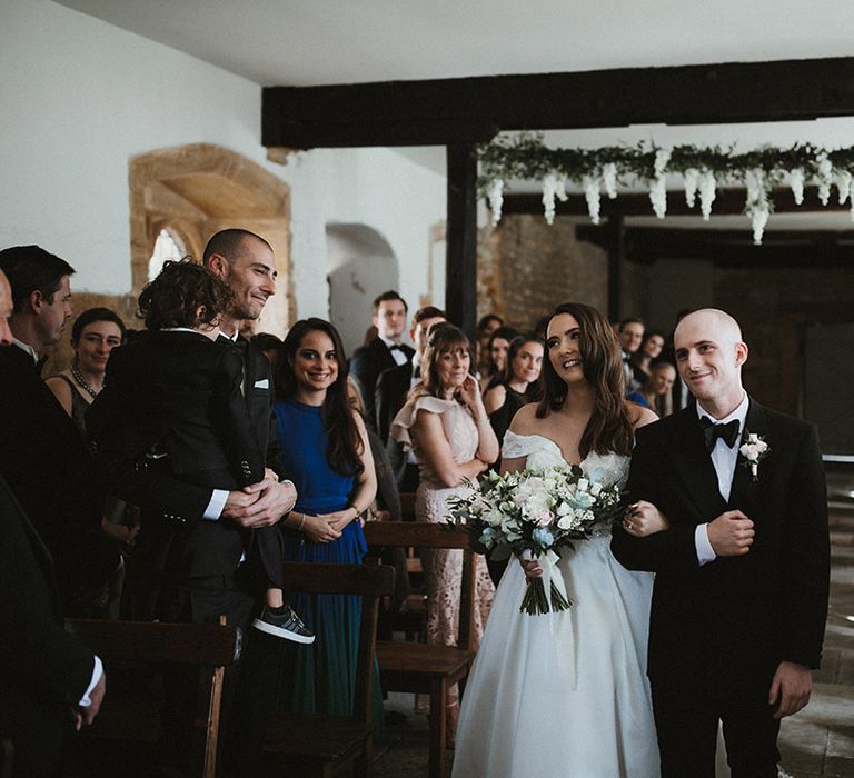 Bride in Pronovias wedding dress walking down the aisle with her brother in black tie 