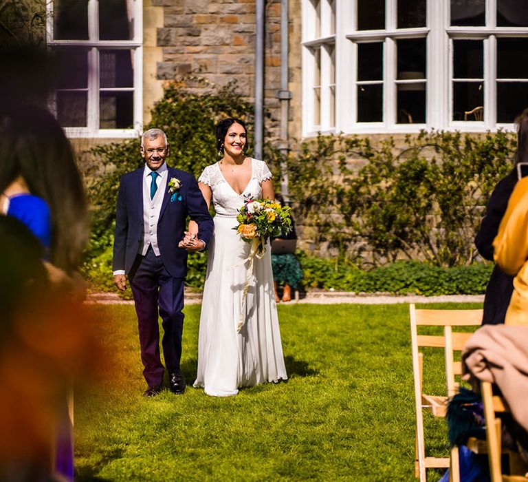 Bride wears lace wedding dress and holds colourful bouquet as she walks down the aisle for outdoor wedding ceremony 