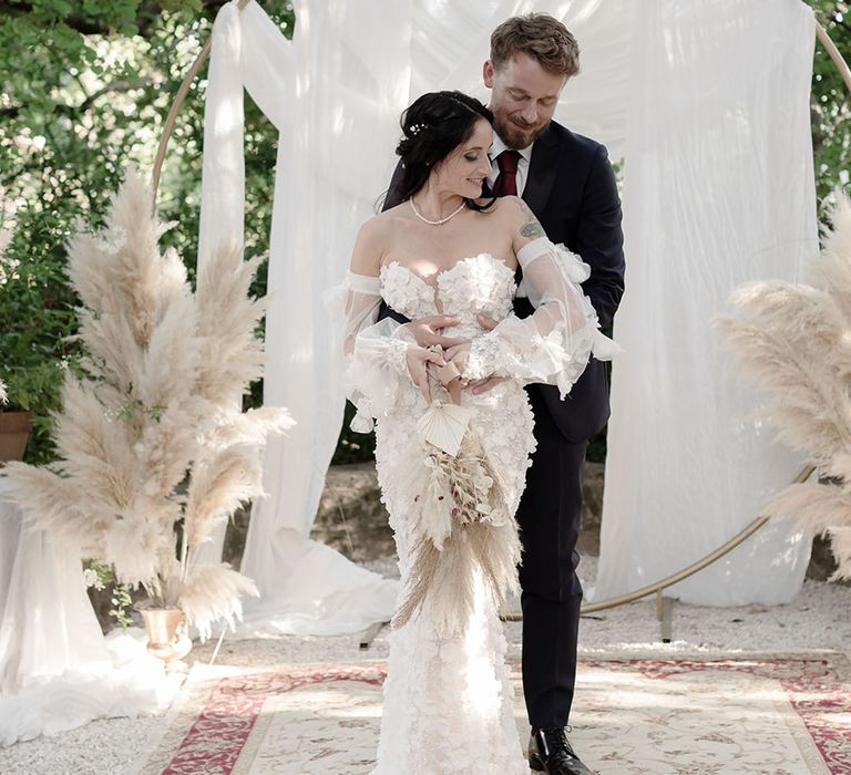 Bride in Pronovias wedding dress holding pampas grass bouquet stands alongside her groom in front of white drapes and pampas grass installations 