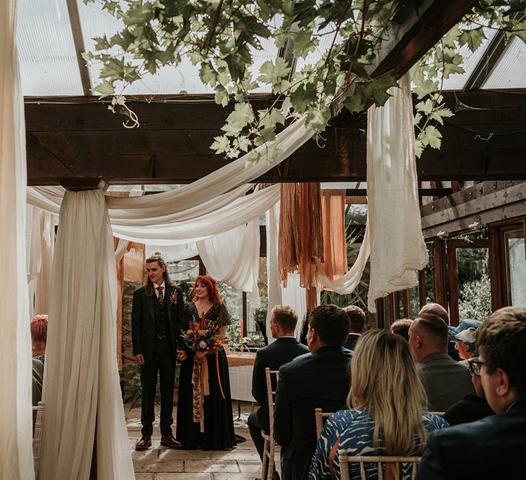 Groom in dark grey three piece suit and burgundy flower patterned tie and bride in embellished black wedding dress holding large orange dried flower bouquet standing amidst off-white, white and orange wedding ceiling drapes decor 