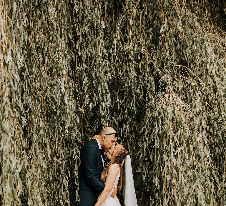 The groom with sunglasses on his head leans in and kisses the bride amongst the leaves 