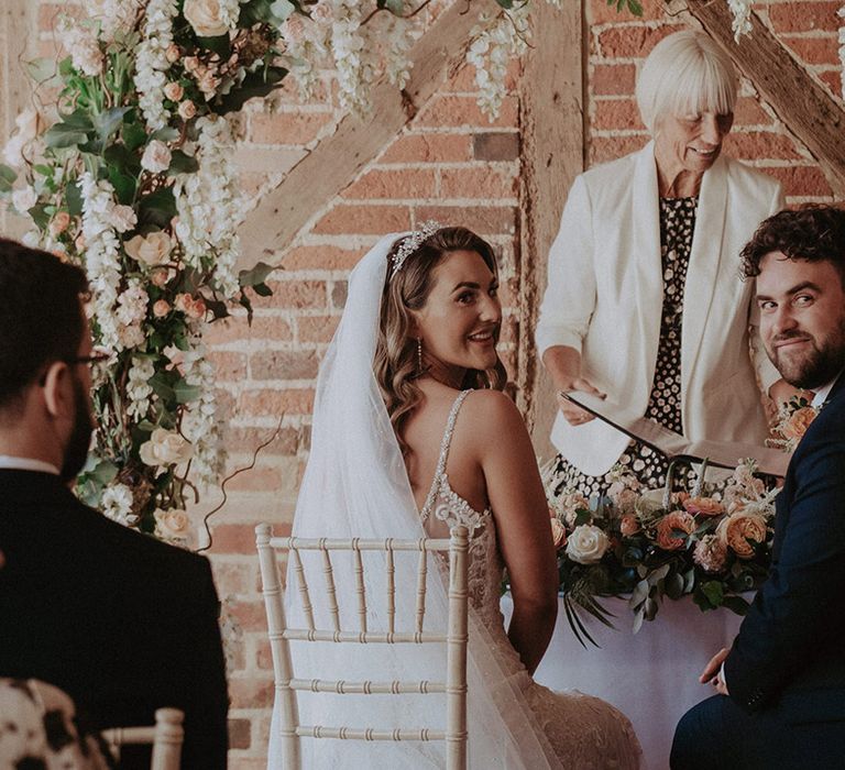 The bride in an embellished wedding dress with tiara and veil sits with the groom in a navy suit as they sign the wedding register 