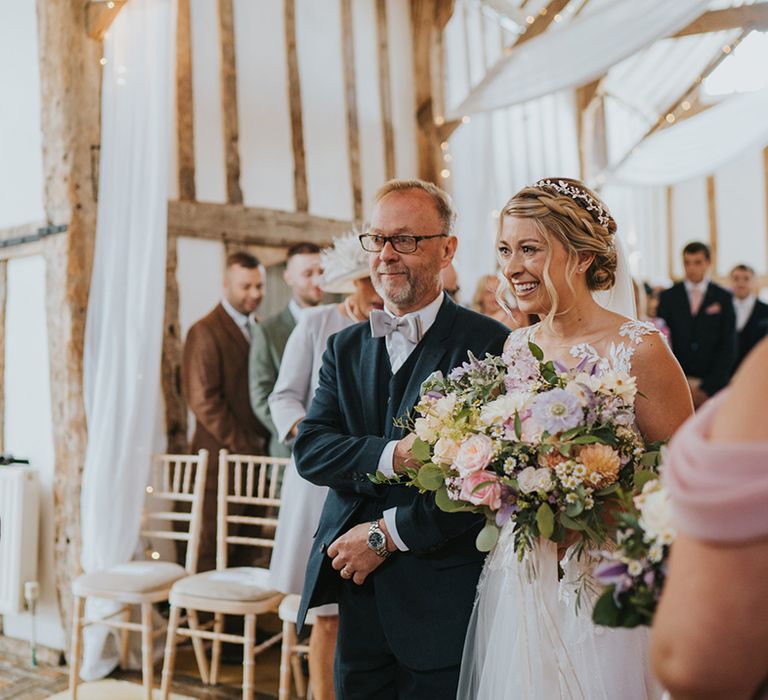 Father of the bride in navy suit with grey bow tie walking bride in illusion lace Dando London wedding dress to the groom holding their son 