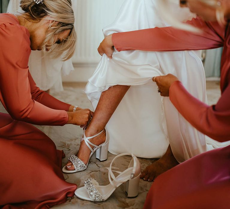 The bridesmaids in orange satin bridesmaid dresses help the bride into her white block heel embellished shoes 