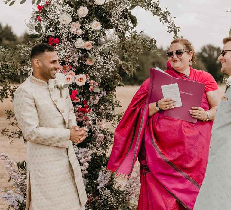 Grooms in traditional wedding sherwanis in the midst of Gujarati wedding ceremony at High Billinghurst Farm wedding venue standing in front of large garden rose, baby's-breath and foliage floral columns