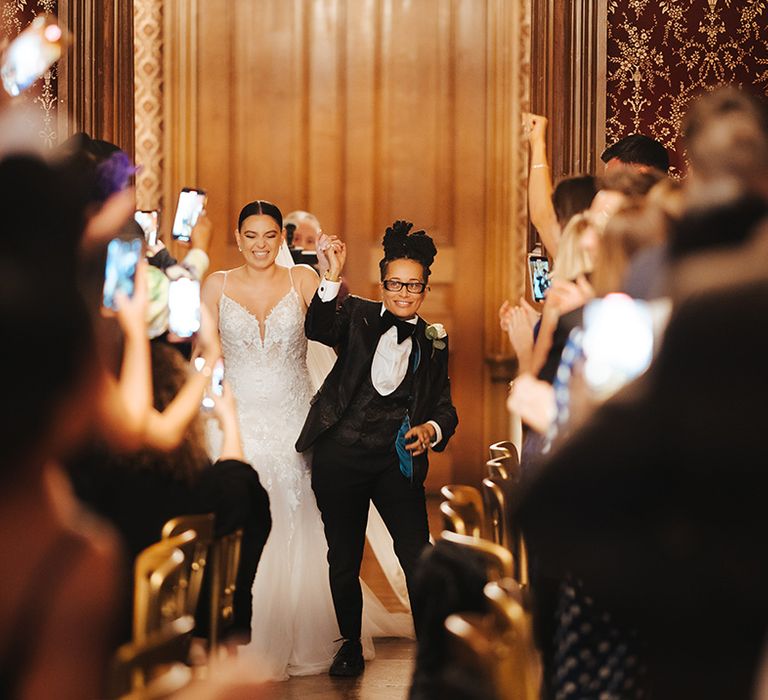 Bride in glasses, black snakeskin print tuxedo with teal lining, black waistcoat, black bowtie and white rose boutonniere walking into the reception room of Grittleton House with bride in lace sleeveless v neck Enzoani wedding dress with tulle finish 