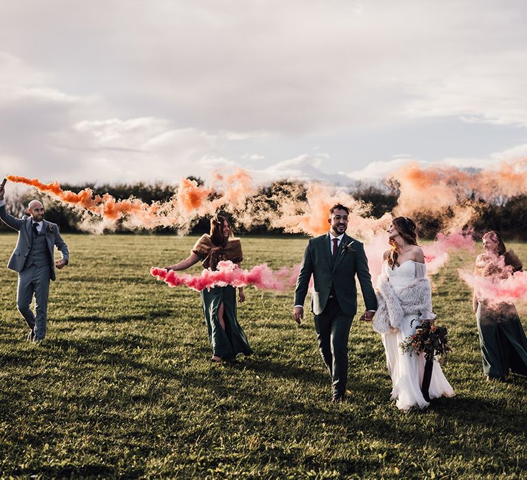 The bridesmaids and groomsmen hold orange and pink wedding smoke bombs with the bride and groom walking together 