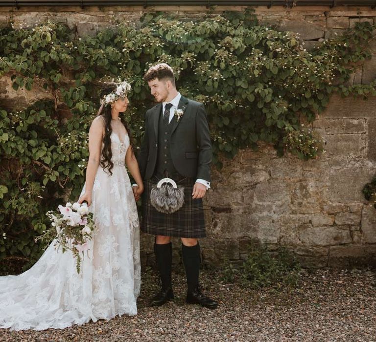 Bride wearing celestial 3d appliqué sheer overlay wedding dress and bridal flower crown holding large white rose, peony, dried flower and foliage bridal bouquet holding hands with groom in grey blazer and waistcoat, dark tartan tie with white flower boutonniere, dark grey and blue tartan kilt and wedding sporran 