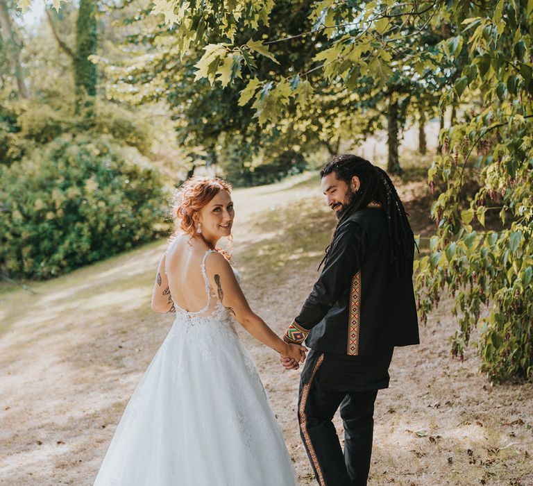 mixed Caribbean groom in a black outfit holding hands with his bride in a white lace wedding dress