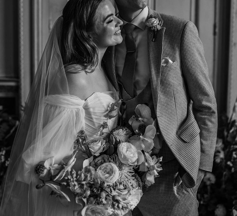 Groom embracing the bride in a houndstooth wedding suit with the bride in an off the shoulder puff sleeve whimsical wedding dress 