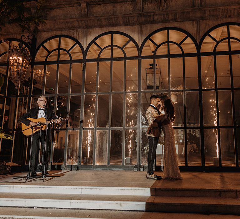 Bride in sparkly off the shoulder reception dress dancing with groom in gold blazer with black lining, black bowtie and black suit trousers on stage with live acoustic guitar player 