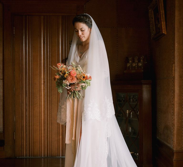 Bride wearing an embroidered edge wedding veil holding orange and red wedding bouquet 