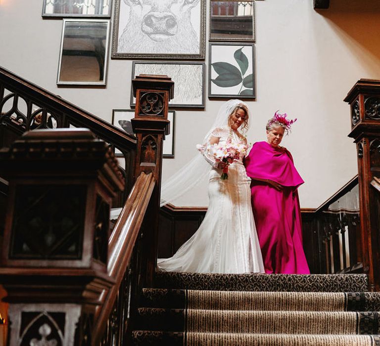 Mother of the bride in bright pink wedding outfit walking down the stairs with the bride in a delicate lace wedding dress 