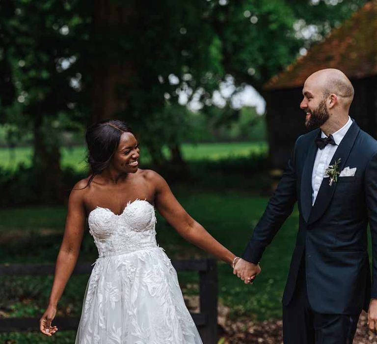 Groom in classic black three piece grooms suit with bow tie and white rose and foliage boutonniere in church holding hands with bride in sweetheart neckline lace wedding dress walking through the grounds after wedding breakfast