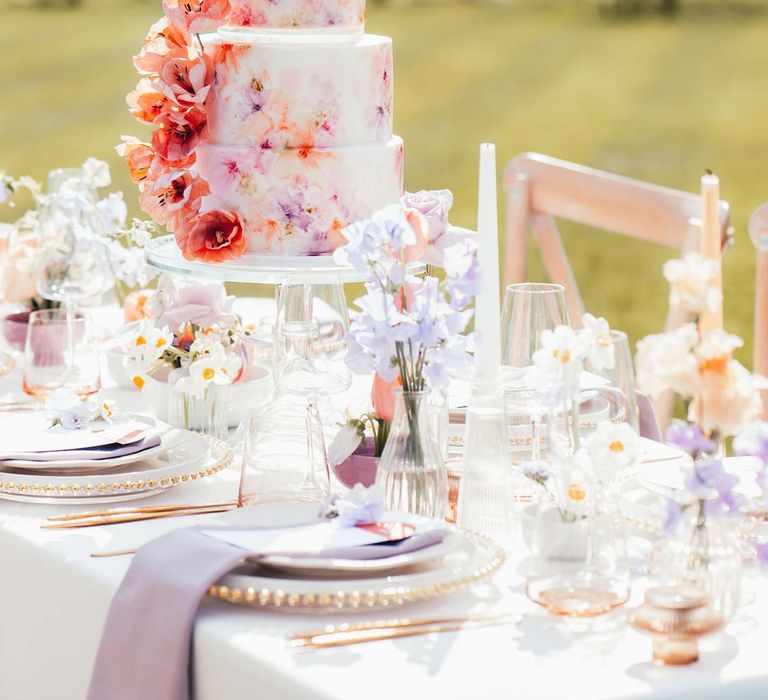 Four tiered hand-painted iced wedding cake on peach and lilac wedding tablescape with spring flower centrepieces, lilac napkins, tinted glassware and gold cutlery and crockery on white tablecloth at Rackleys Barn