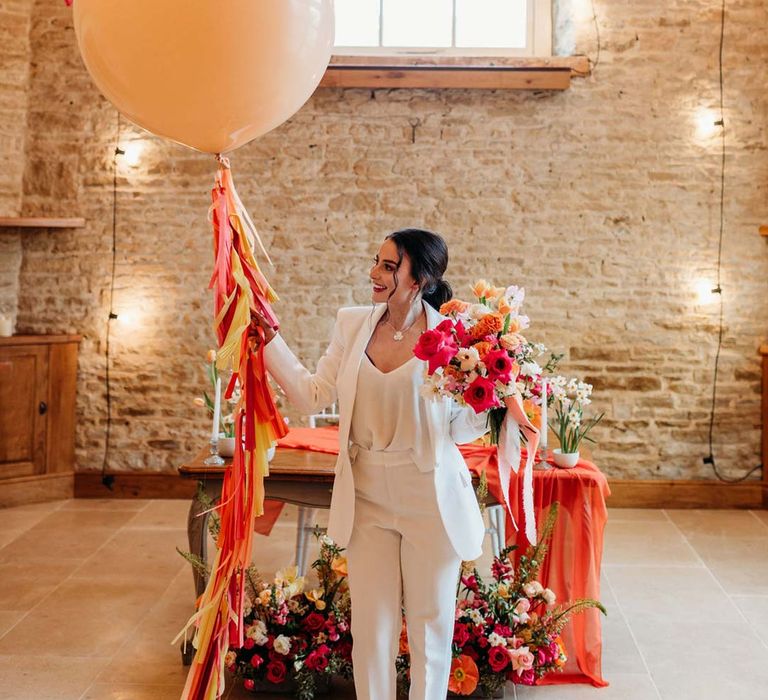 Bride in white bridal suit in Merriscourt wedding venue reception room with metallic pink, white, orange and yellow balloons, wedding fairy lights decorations and wooden table with orange table runner and mixed seasonal flower arrangements 