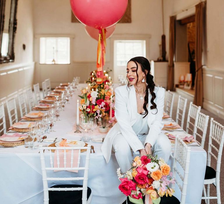 Bride in white bridal suit with silver bridal jewellery with closed toe bright green wedding heels holding colourful bridal bouquet sitting on eclectic colourful wedding tablescape at Merriscourt wedding venue